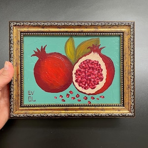 Könnte beinhalten: Gerahmtes Gemälde mit zwei Granatäpfeln, einem ganzen und einem halbierten, die die Samen zeigen. Das Kunstwerk hat einen türkisfarbenen Hintergrund und einen goldfarbenen, verzierten Rahmen. Die Granatäpfel sind in Rottönen mit grünen Blättern gemalt.
