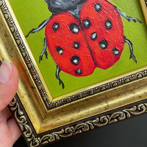 May include: A framed painting of a ladybug. The ladybug is red with black spots and a black head, set against a green background. The frame is ornate, with gold and brown tones.