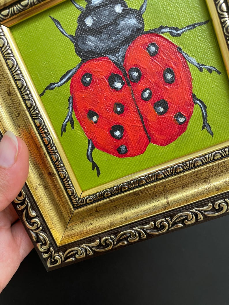 May include: A framed painting of a ladybug. The ladybug is red with black spots and a black head, set against a green background. The frame is ornate, with gold and brown tones.