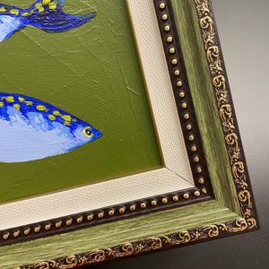 May include: An original framed painting featuring two blue fish with yellow spots against a green background. The frame is green with gold accents and a cream-colored inner border. The artwork is a close-up view.