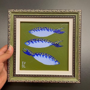 May include: An original painting of three blue fish with yellow accents on a green background. The artwork is framed in a decorative green and gold frame. The artist's initials, EV OL, are visible in the lower left corner.