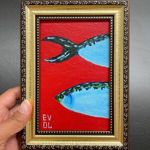 May include: A framed painting of two blue fish on a red background. The fish have green and black details. The frame is gold with a decorative border. The artist's initials are in the lower left corner.