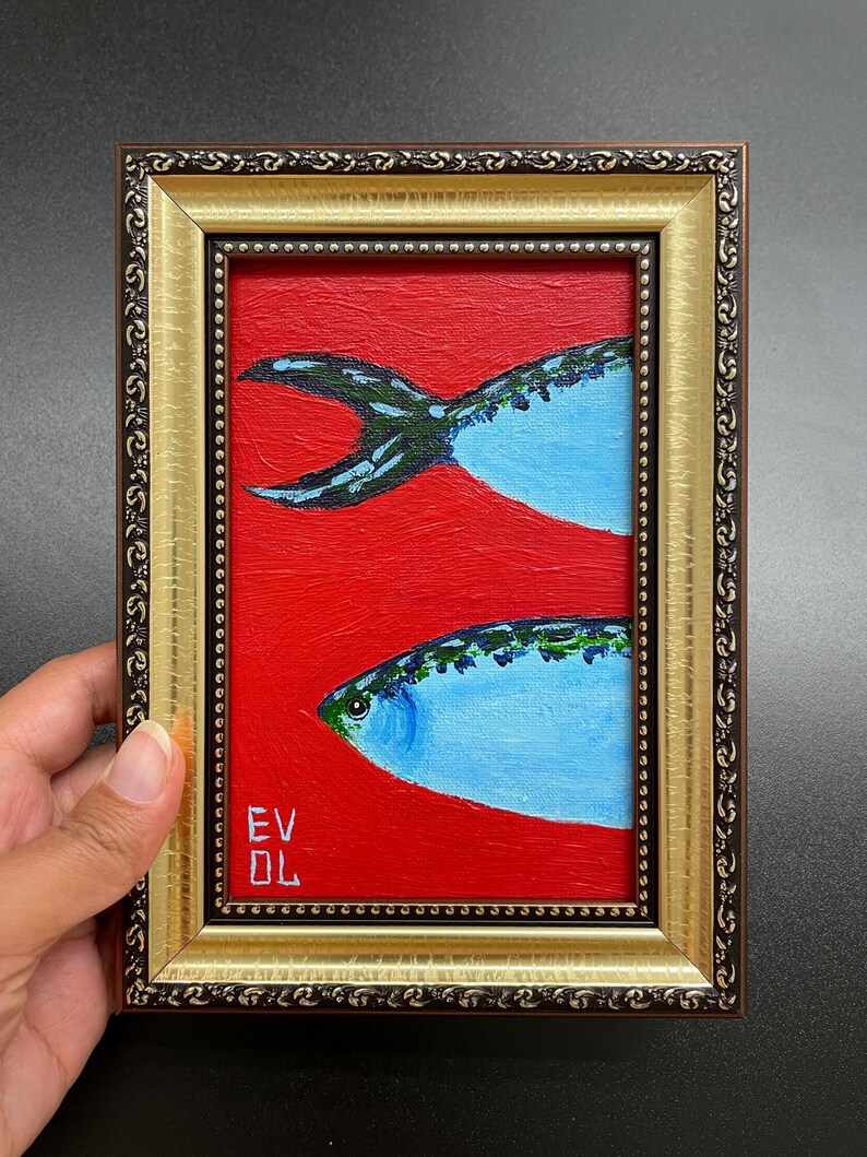 May include: A framed painting of two blue fish on a red background. The fish have green and black details. The frame is gold with a decorative border. The artist's initials are in the lower left corner.