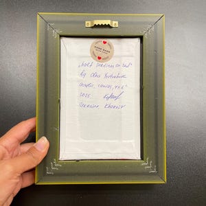 May include: Back of a framed artwork with a green and yellow frame. The artwork is labeled "half sardines on red" by Olha Yeutukhova, acrylic on canvas, 4x6 inches. Includes a "Hand Made With Love" sticker and handwritten text.