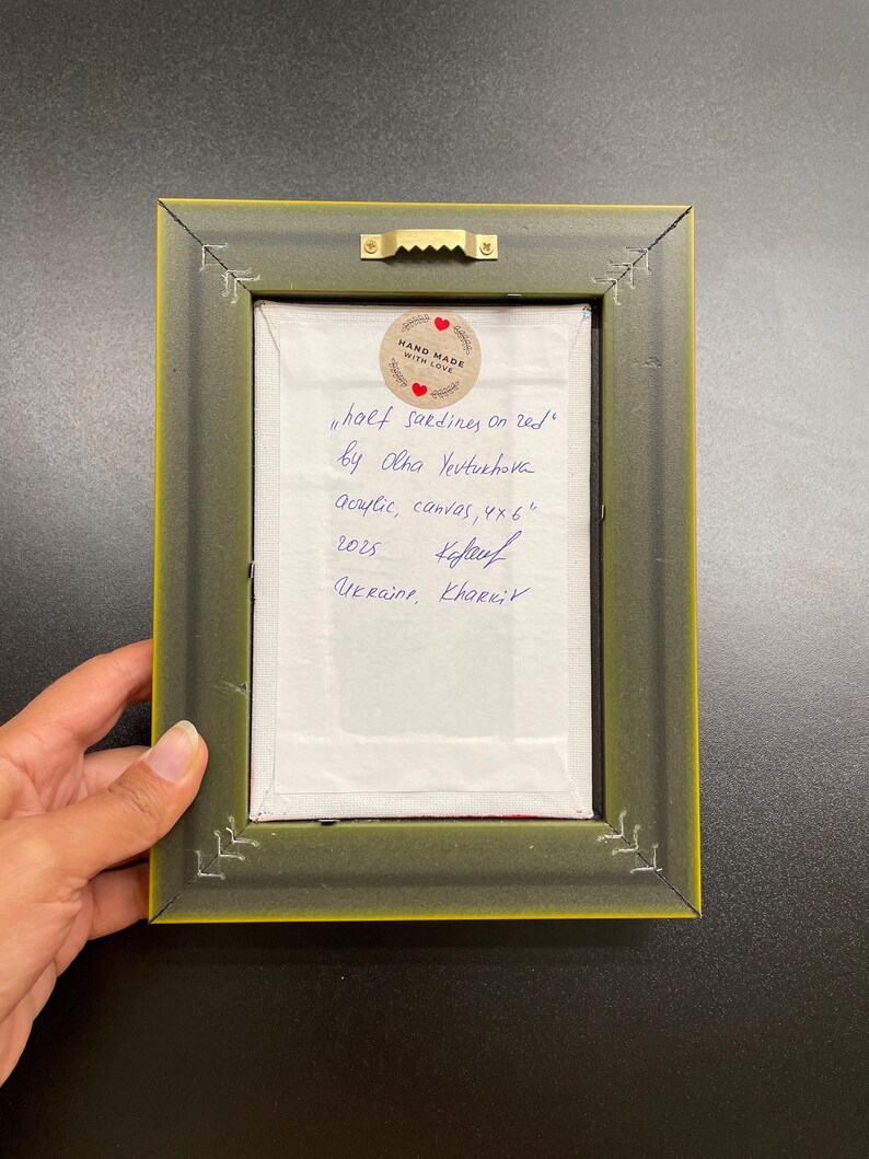 May include: Back of a framed artwork with a green and yellow frame. The artwork is labeled "half sardines on red" by Olha Yeutukhova, acrylic on canvas, 4x6 inches. Includes a "Hand Made With Love" sticker and handwritten text.