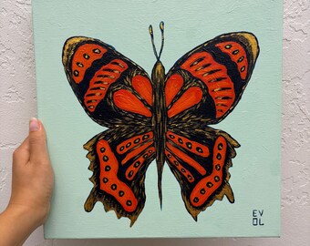 May include: A square canvas painting featuring a butterfly with vibrant orange and black wings, accented with gold outlines, set against a light mint green background. The text EV OL is printed in the bottom right corner of the canvas.