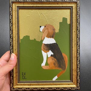 Könnte beinhalten: Gerahmtes Gemälde eines Beagle-Hundes in sitzender Position. Der Hund ist braun, schwarz und weiß, vor einem grünen Hintergrund mit goldenen Akzenten. Der Rahmen ist mit einer goldenen Oberfläche verziert. Die Initialen des Künstlers "EV OL" befinden sich in der unteren linken Ecke.