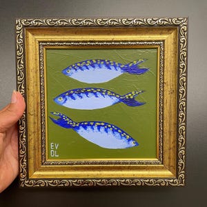May include: A framed painting featuring three blue fish with yellow accents on a green background. The fish are painted with a light blue body and dark blue fins. The frame is gold with ornate detailing. The artist's initials are in the lower left corner.
