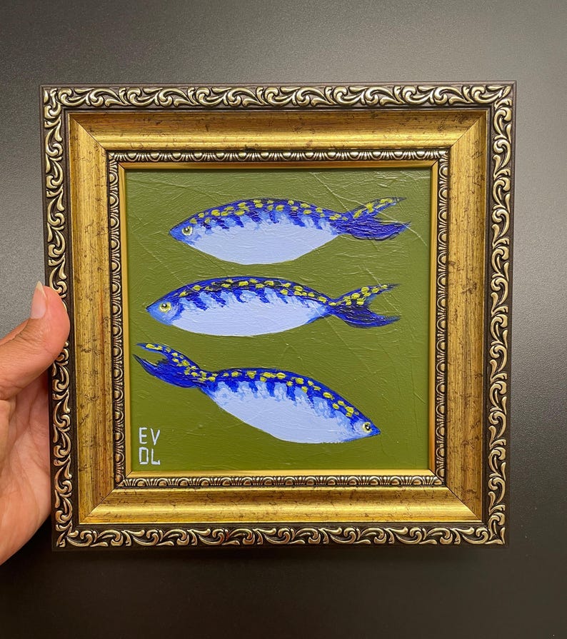 May include: A framed painting featuring three blue fish with yellow accents on a green background. The fish are painted with a light blue body and dark blue fins. The frame is gold with ornate detailing. The artist's initials are in the lower left corner.