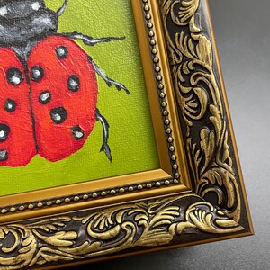 May include: A framed painting of a ladybug. The ladybug is red with black spots and a black head, set against a green background. The ornate gold frame has a dark brown border with detailed carvings.