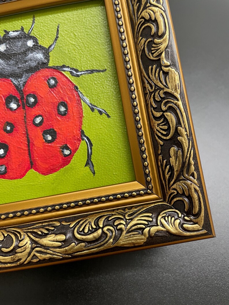 May include: A framed painting of a ladybug. The ladybug is red with black spots and a black head, set against a green background. The ornate gold frame has a dark brown border with detailed carvings.