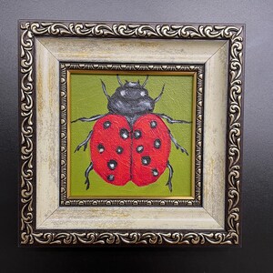 May include: A framed painting of a ladybug. The ladybug is red with black spots and a black head, set against a green background. The frame is ornate, with a cream and brown finish.