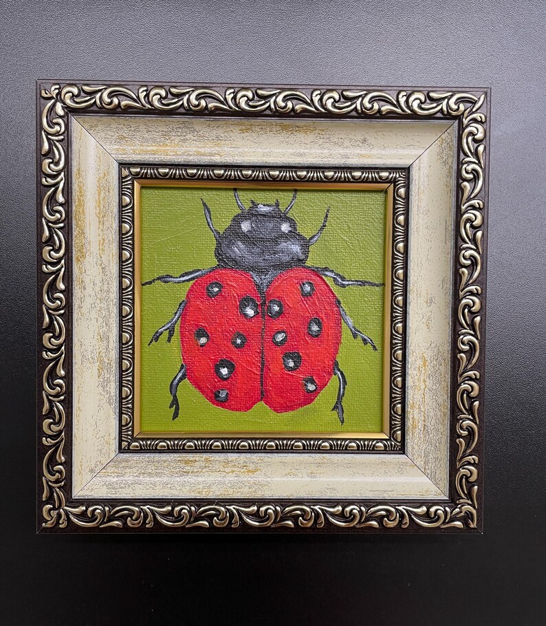 May include: A framed painting of a ladybug. The ladybug is red with black spots and a black head, set against a green background. The frame is ornate, with a cream and brown finish.