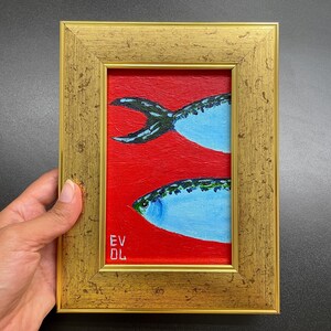 May include: A framed painting featuring two stylized fish against a red background. The fish are light blue with green and black details, and the frame is gold. The artist's initials, EV DL, are visible in the lower left corner.