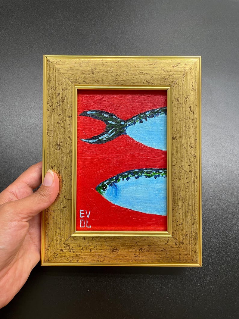 May include: A framed painting featuring two stylized fish against a red background. The fish are light blue with green and black details, and the frame is gold. The artist's initials, EV DL, are visible in the lower left corner.