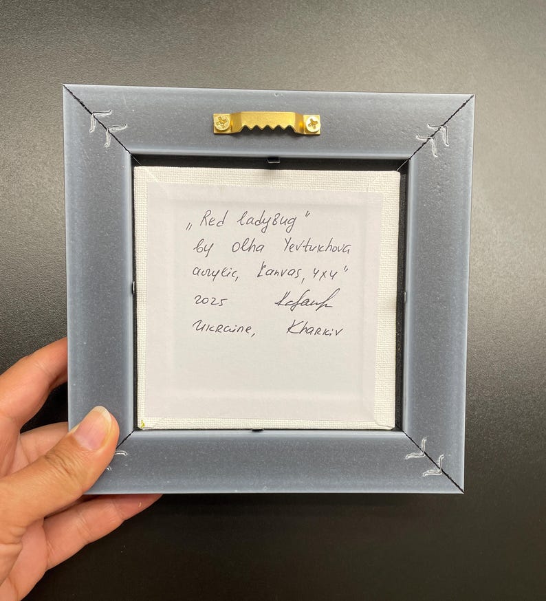 May include: Back of a framed artwork titled "Red ladyBug" by Olha Yevtukhova. The artwork is acrylic on canvas, measuring 4x4 inches. The frame is gray with decorative corner accents. The text includes the year 2025 and the location Ukraine, Kharkiv.