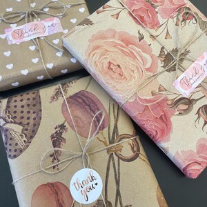 May include: Wrapped gifts with decorative paper and twine. One gift is wrapped in brown paper with white hearts, another with pink roses, and a third with macarons and lavender. Each gift has a "Thank you" sticker.