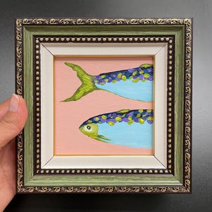 以下が含まれることがあります： 2匹の様式化された魚を描いた、額装された小さなアートワーク。魚は青い体と紫と黄色のディテールがあり、ピンクの背景に配置されています。フレームは緑色で、金色のアクセントが付いています。
