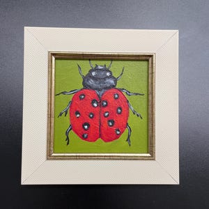 May include: A framed painting of a ladybug. The ladybug has a red shell with black spots and a black head. The background is green, and the frame is off-white with a gold inner edge. The artwork is square.