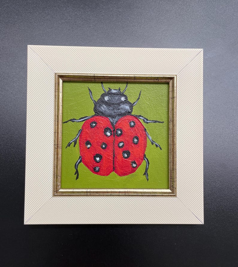 May include: A framed painting of a ladybug. The ladybug has a red shell with black spots and a black head. The background is green, and the frame is off-white with a gold inner edge. The artwork is square.