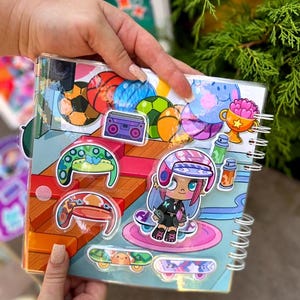 May include: A spiral-bound notebook with colorful stickers featuring sports balls, a boombox, and a cartoon skater girl. The notebook has a clear plastic cover and is held by a person's hands. The stickers are in various colors, including blue, orange, and green.