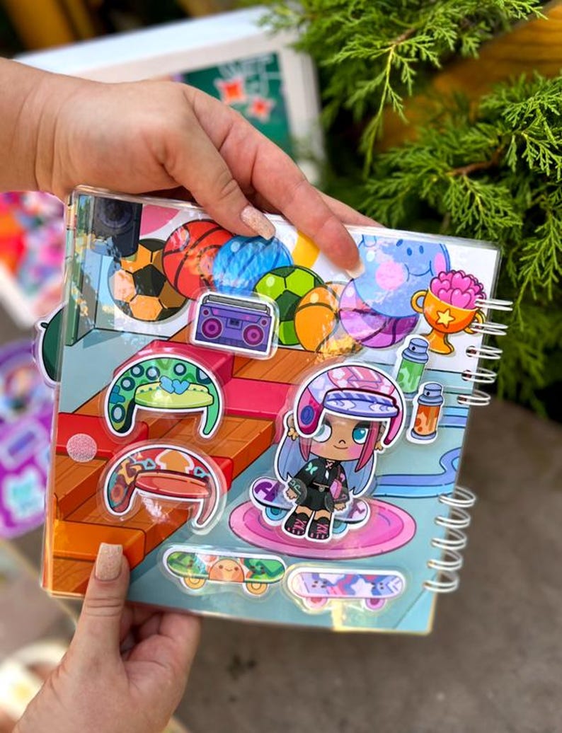 May include: A spiral-bound notebook with colorful stickers featuring sports balls, a boombox, and a cartoon skater girl. The notebook has a clear plastic cover and is held by a person's hands. The stickers are in various colors, including blue, orange, and green.