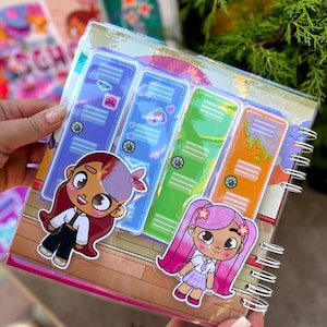 May include: A spiral-bound notebook with a clear plastic cover featuring colorful cartoon locker designs. Two cartoon girl stickers are on the cover. The notebook has a wooden floor design and is held by a person.