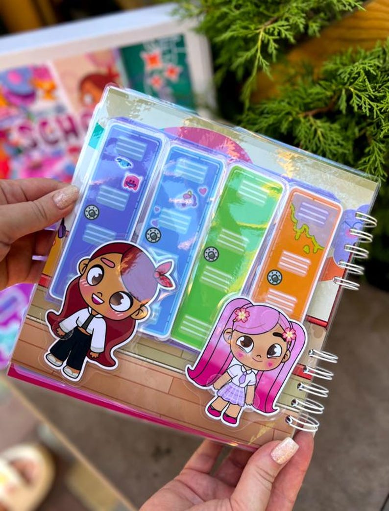 May include: A spiral-bound notebook with a clear plastic cover featuring colorful cartoon locker designs. Two cartoon girl stickers are on the cover. The notebook has a wooden floor design and is held by a person.