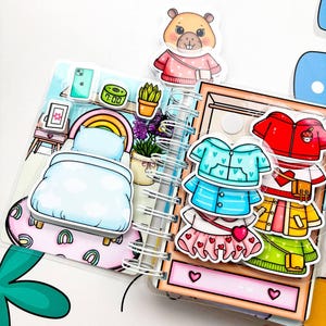 May include: A spiral-bound paper doll book with a bedroom scene and clothing options. The bedroom scene includes a bed, rainbow, and potted plant. Clothing options include a blue jacket, red jacket, and skirt. A cartoon beaver is at the top.