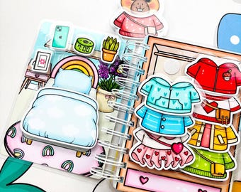 May include: A spiral-bound paper doll book with a bedroom scene and clothing options. The bedroom scene includes a bed, rainbow, and potted plant. Clothing options include a blue jacket, red jacket, and skirt. A cartoon beaver is at the top.