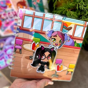 May include: A spiral-bound notebook featuring colorful cartoon illustrations of characters in a diner setting. The cover has a clear plastic overlay with stickers of two cartoon girls. The background includes tables, chairs, and condiments.