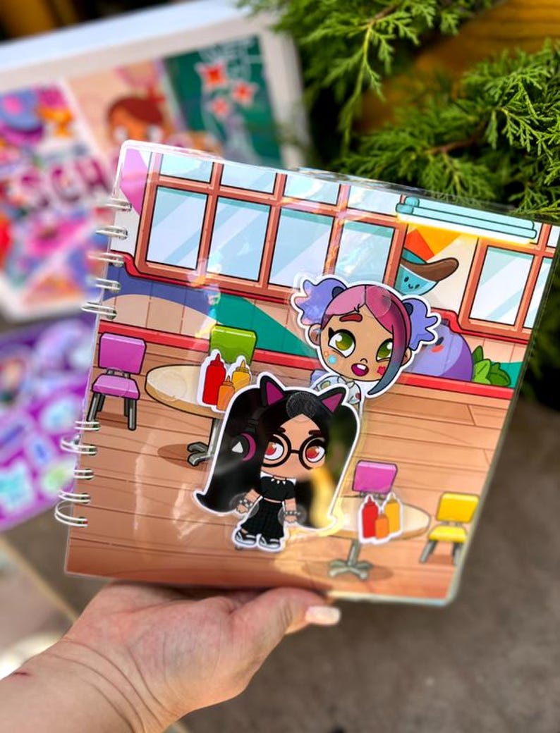 May include: A spiral-bound notebook featuring colorful cartoon illustrations of characters in a diner setting. The cover has a clear plastic overlay with stickers of two cartoon girls. The background includes tables, chairs, and condiments.