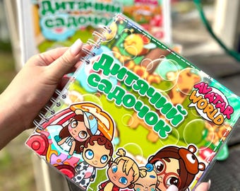 May include: A spiral-bound children's book with the text "ДитЯчий садочок" and cartoon illustrations. The cover features colorful characters and the words "Avatar World". The book is held in front of a blurred background.