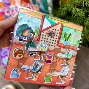 May include: A spiral-bound sticker book featuring a school-themed design. The cover shows a cartoon student and classroom scene. Stickers include backpacks, desks, pencils, and a dinosaur. The color palette includes orange, green, blue, and brown.