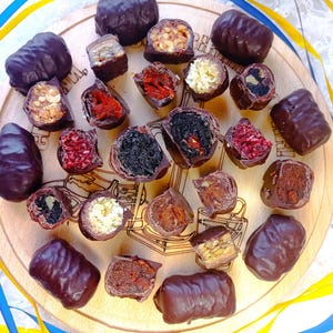 May include: An assortment of chocolate-covered candies with various fillings, including nuts, dried fruits, and creamy centers, arranged on a wooden serving board. The candies are dark brown, with visible textures and fillings. Yellow and blue ribbons are in the background.
