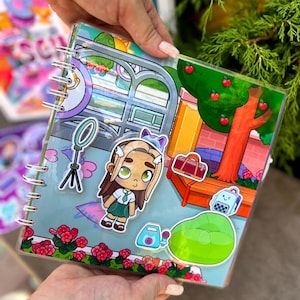 May include: A clear, spiral-bound notebook with a colorful cartoon illustration. The illustration features a girl, a tree with red apples, a beanbag chair, and other accessories. The notebook is held by a person.