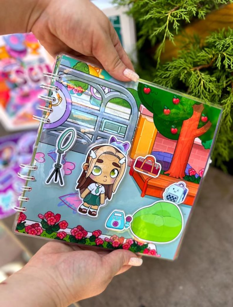 May include: A clear, spiral-bound notebook with a colorful cartoon illustration. The illustration features a girl, a tree with red apples, a beanbag chair, and other accessories. The notebook is held by a person.