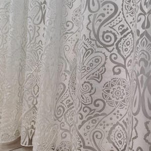 Op de afbeelding: Witte, doorschijnende gordijn met een grijs paisley-patroon. Het gordijn is gemaakt van een lichtgewicht stof en heeft een delicaat, vloeiend ontwerp.