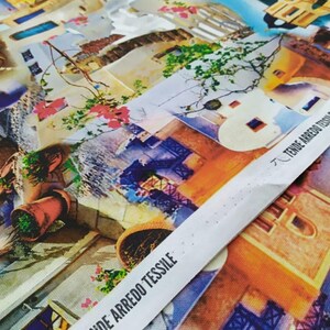 Può includere: Un tessuto colorato con una stampa di edifici e piante in stile acquerello. Il tessuto ha un'etichetta bianca con il testo "Tende Arredo Tessile" in nero.
