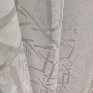 Può includere: Primo piano di tessuto bianco con un motivo sottile e ripetuto in grigio chiaro. Il motivo sembra essere un disegno astratto di linee e forme. Il tessuto è drappeggiato, creando pieghe e ombre morbide. L'effetto complessivo è delicato e strutturato.