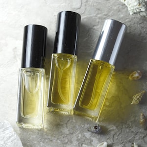 Pode incluir: Três pequenos frascos de perfume retangulares com tampas pretas e prateadas, cheios de um líquido amarelo. Os frascos estão dispostos sobre uma superfície cinza claro, com pequenas conchas marinhas espalhadas.