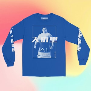 Może przedstawiać: Długi rękaw t-shirt w kolorze królewskiego błękitu z białym nadrukiem przedstawiającym zapaśnika sumo. Nadruk zawiera tekst "大鵬" oraz imię zapaśnika w mniejszym rozmiarze czcionki. Rękawy mają biały nadruk przedstawiający kwiat.