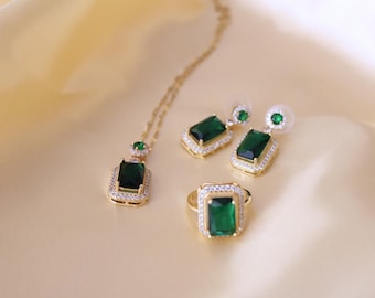 May include: A gold-toned jewelry set featuring a necklace, earrings, and a ring. Each piece showcases a large, emerald-green rectangular gemstone surrounded by small, clear stones. The set is displayed on a cream-colored, satin-like surface.