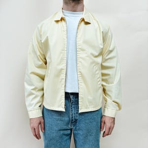 May include: Pale yellow jacket with a collared neckline and a zip closure. The jacket has long sleeves and a relaxed fit. The person is wearing a white t-shirt and blue jeans. The jacket is a light, neutral colour.