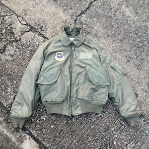 May include: Olive green bomber jacket with a zip-up front, two front pockets, and a patch on the chest. The jacket has a ribbed collar, cuffs, and hem. A small patch is visible on the left shoulder. The jacket is made of a shiny material.