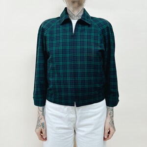 May include: A green and navy blue plaid jacket with a zip-up front and a collared neckline. The jacket has long sleeves and is paired with white pants. The jacket's plaid pattern is a classic design.