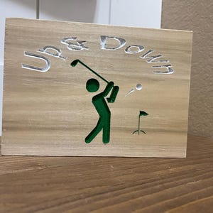 Pode incluir: Placa de madeira com as palavras "Up & Down" gravadas no topo. Uma silhueta verde de um jogador de golfe a balançar um taco, com uma bola de golfe no ar. Uma pequena bandeira também é esculpida na madeira.
