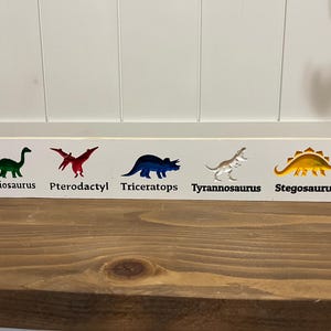 Pode incluir: Uma placa de madeira branca com silhuetas coloridas de dinossauros: Brachiosaurus, Pterodáctilo, Triceratops, Tyrannosaurus e Stegosaurus. Cada dinossauro é rotulado com seu nome em texto preto. A placa está em uma superfície de madeira.