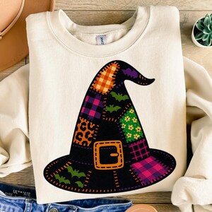 Puede incluir: Sudadera color crema con un diseño de sombrero de bruja grande y colorido. El sombrero es negro con patrones de patchwork en naranja, morado, verde y rosa, incluyendo murciélagos, flores y cuadros. Una hebilla naranja acentúa la base del sombrero, ideal para Halloween.