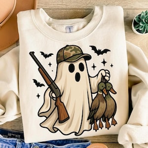 May include: Cream-colored sweatshirt featuring a cartoon ghost wearing a camouflage hat and holding a shotgun and two ducks. Black bats and stars surround the ghost. The design is printed on a soft, comfortable fabric.